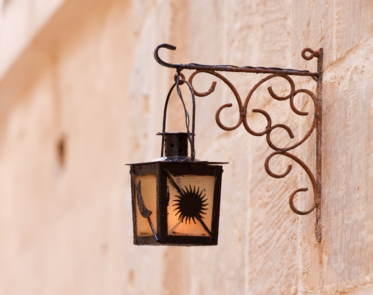 Applique lanterne ancienne en fer forgé noir sur mur en pierre claire, motif soleil et lumière ambrée